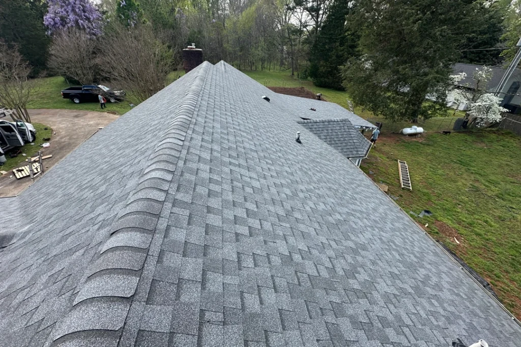 High-angle view of a large, newly installed gray shingle roof featuring clean ridge caps, multiple roof vents, and uniform shingle alignment, with workers, vehicles, and a wooded backyard visible below.