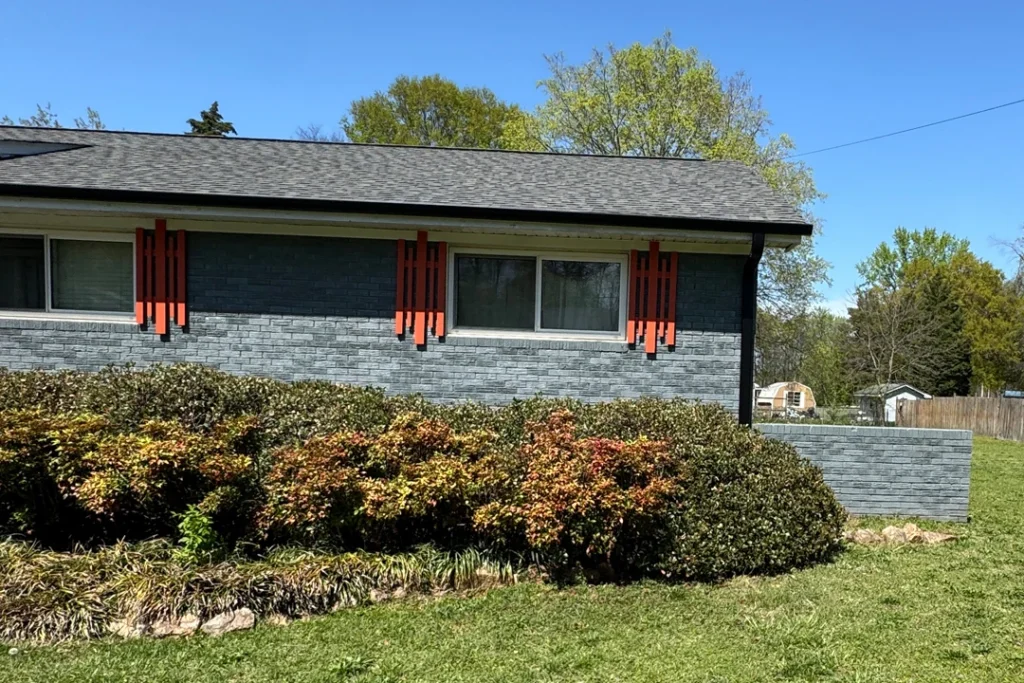 Front view of a single story brick home with gray painted exterior, red decorative shutters, a dark shingle roof, and neatly trimmed bushes in the yard.