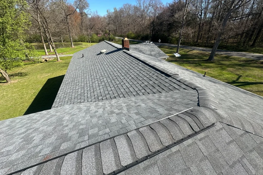 New gray asphalt shingle roof with ridge venting on a residential home, showing clean installation lines and surrounding yard and trees on a sunny day.