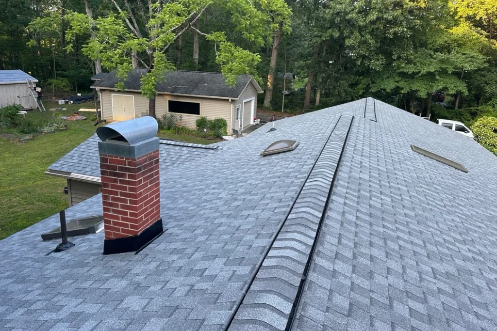 A rooftop view showing new gray shingles, ridge vents, a brick chimney, and skylights on a residential home, with a yard and detached garage in the background.
